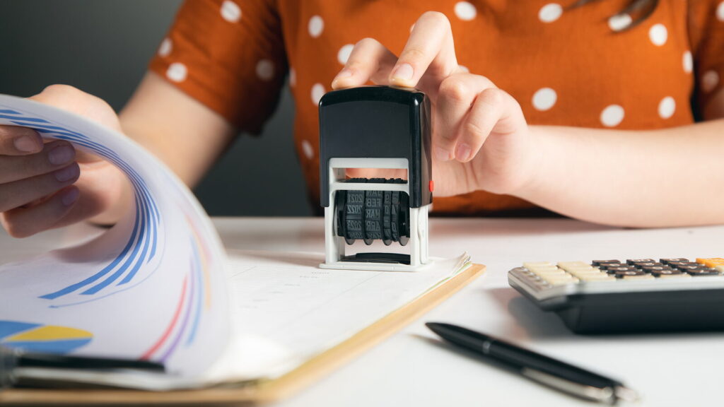 Mulher carimba um documento ao lado de uma calculadora, o custo de uma apostila