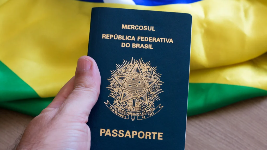 Mão segura um passaporte brasileiro diante da bandeira do Brasil